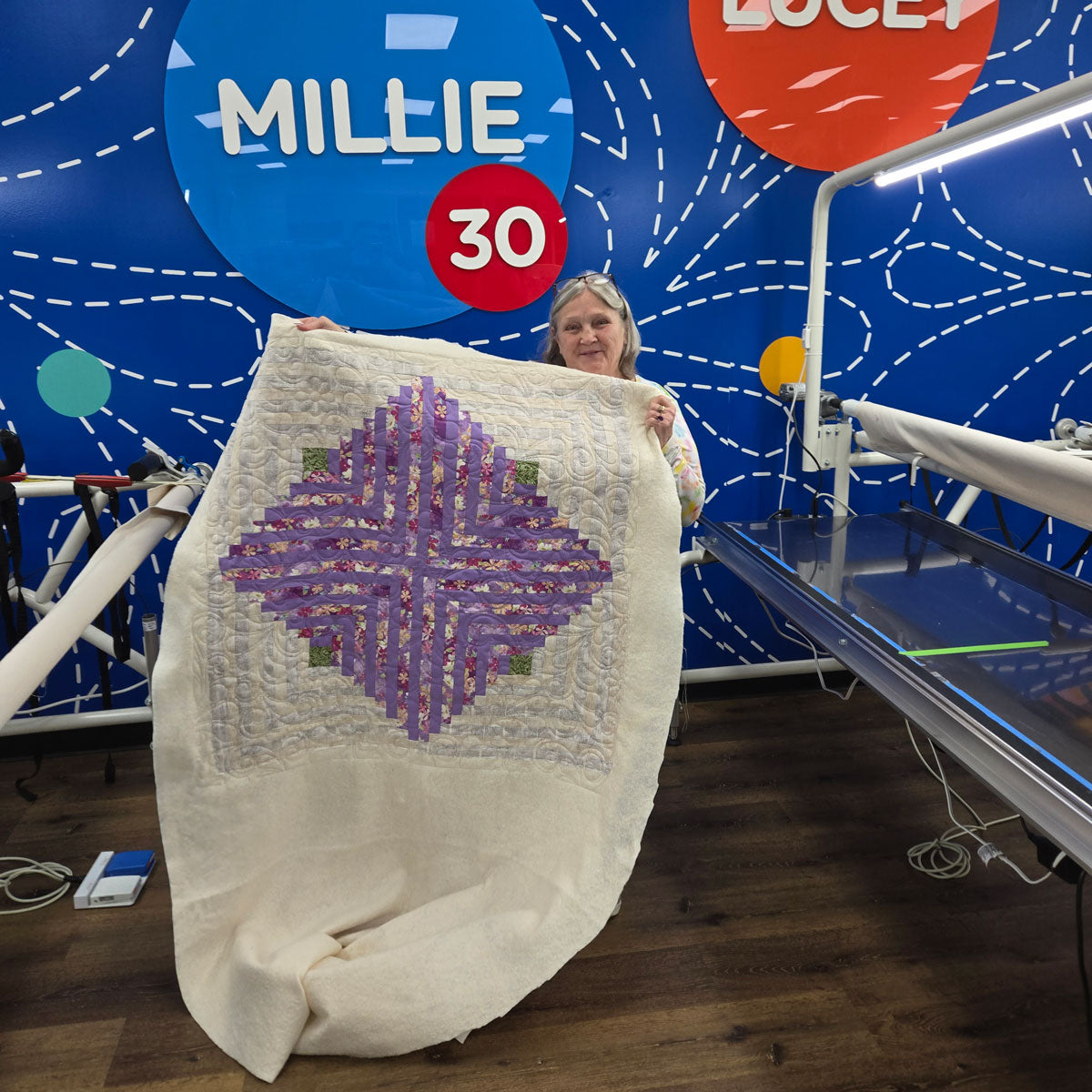 A woman stands indoors holding a large quilt with a purple geometric pattern. Behind her is a blue wall with colorful signs reading MILLIE 30 and LUCEY, along with quilting machines and equipment visible.