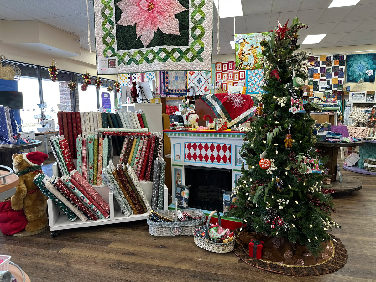A cozy quilt shop decorated for Christmas with fabric bolts, a holiday quilt, a faux fireplace, baskets of thread, and a decorated Christmas tree with ornaments and presents underneath.