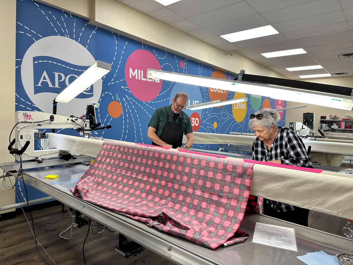 Two people work together at a long quilting machine, arranging a pink and gray checkered fabric. The workspace has bright lighting and colorful wall art with words like APQS and MILE.