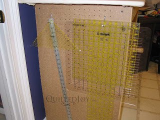 Clear plastic quilting rulers and a metal ruler hanging on hooks on a brown pegboard side of a worktable, with part of a tiled floor and a white door frame visible nearby.