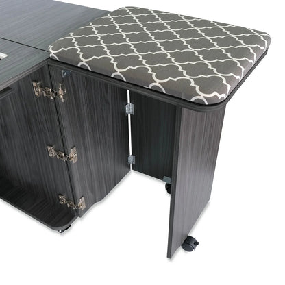 A cushioned stool with a gray and white geometric pattern is partially tucked under the Arrow Companies, LLC Kangaroo Sydney Sewing Cabinet with Electric Lift, which features multiple hinges and caster wheels.