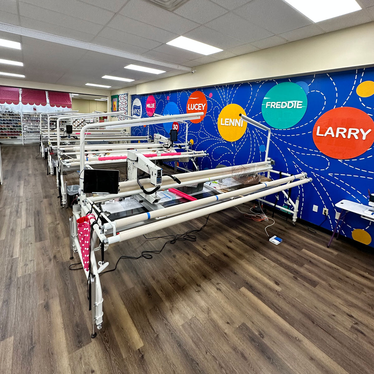 Rows of longarm quilting machines are set up in a bright, spacious room with wood floors. A colorful wall displays names—Allie, Lucey, Lenni, Freddie, and Larry—in large circles.