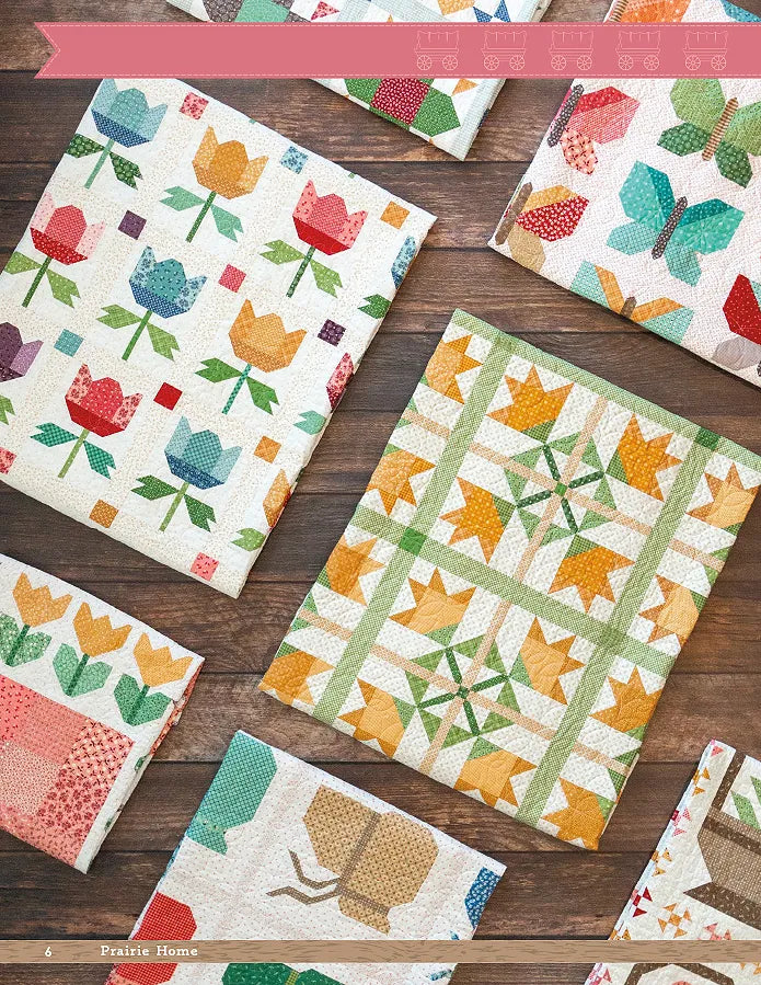 Several vibrant quilts with tulip, butterfly, and patchwork motifs are neatly folded on a wooden surface. Their designs are inspired by the Prairie Home Quilt Book from Checker Distributor, featuring floral and geometric quilting patterns.
