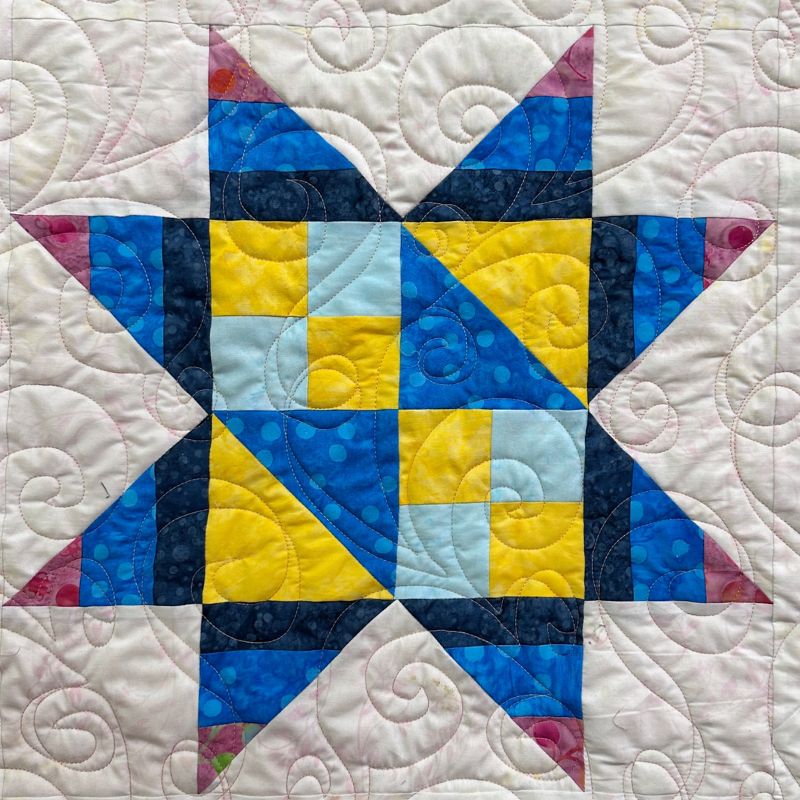 A quilt block with a star pattern made from blue, yellow, and pink fabrics on a white background. The star has eight points and is pieced with various patterned fabrics. Spiral quilt stitching decorates the entire design.
