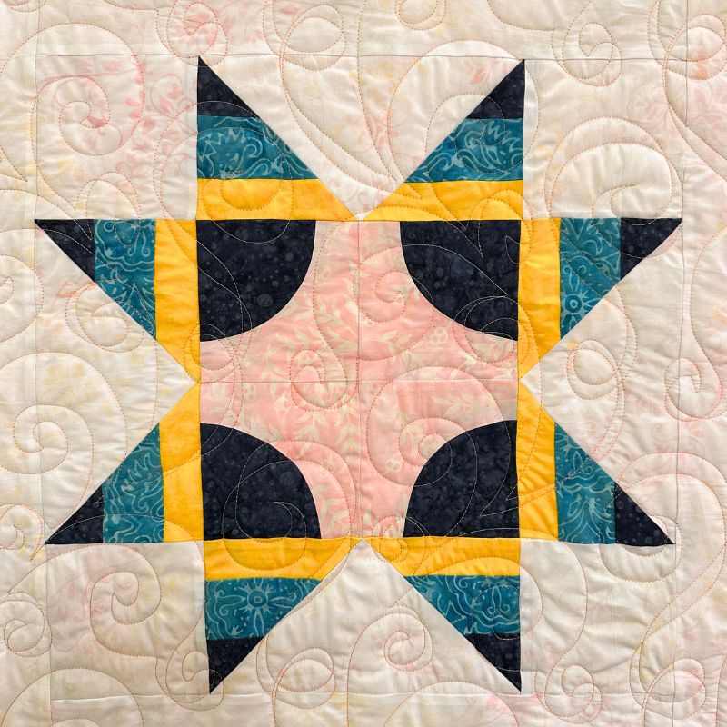 A quilt block featuring a star pattern with dark blue, teal, yellow, and pink fabric pieces on a cream background, accented with swirling quilted stitching throughout.