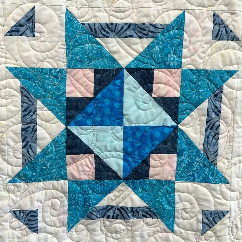 A quilt block featuring a blue and teal star pattern with light pink and white accents, surrounded by intricate swirling stitching on a light background.