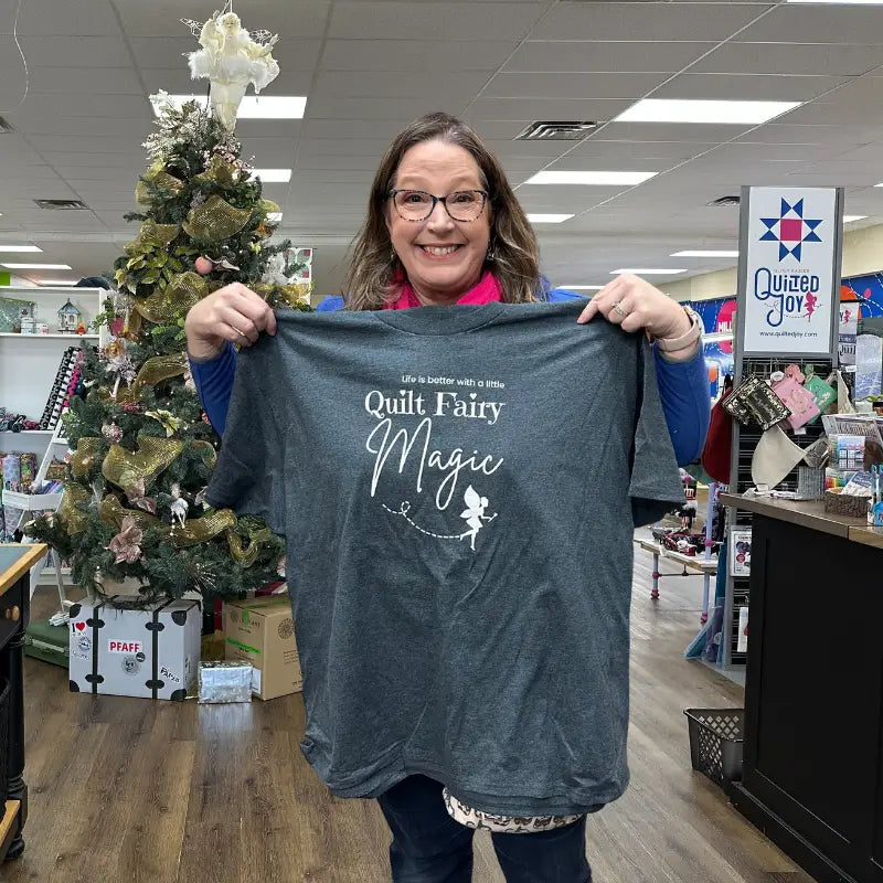A smiling woman stands indoors, holding up a gray T-shirt that reads Life is better with a little Quilt Fairy Magic with a small fairy graphic. A decorated Christmas tree and quilting supplies are visible in the background.
