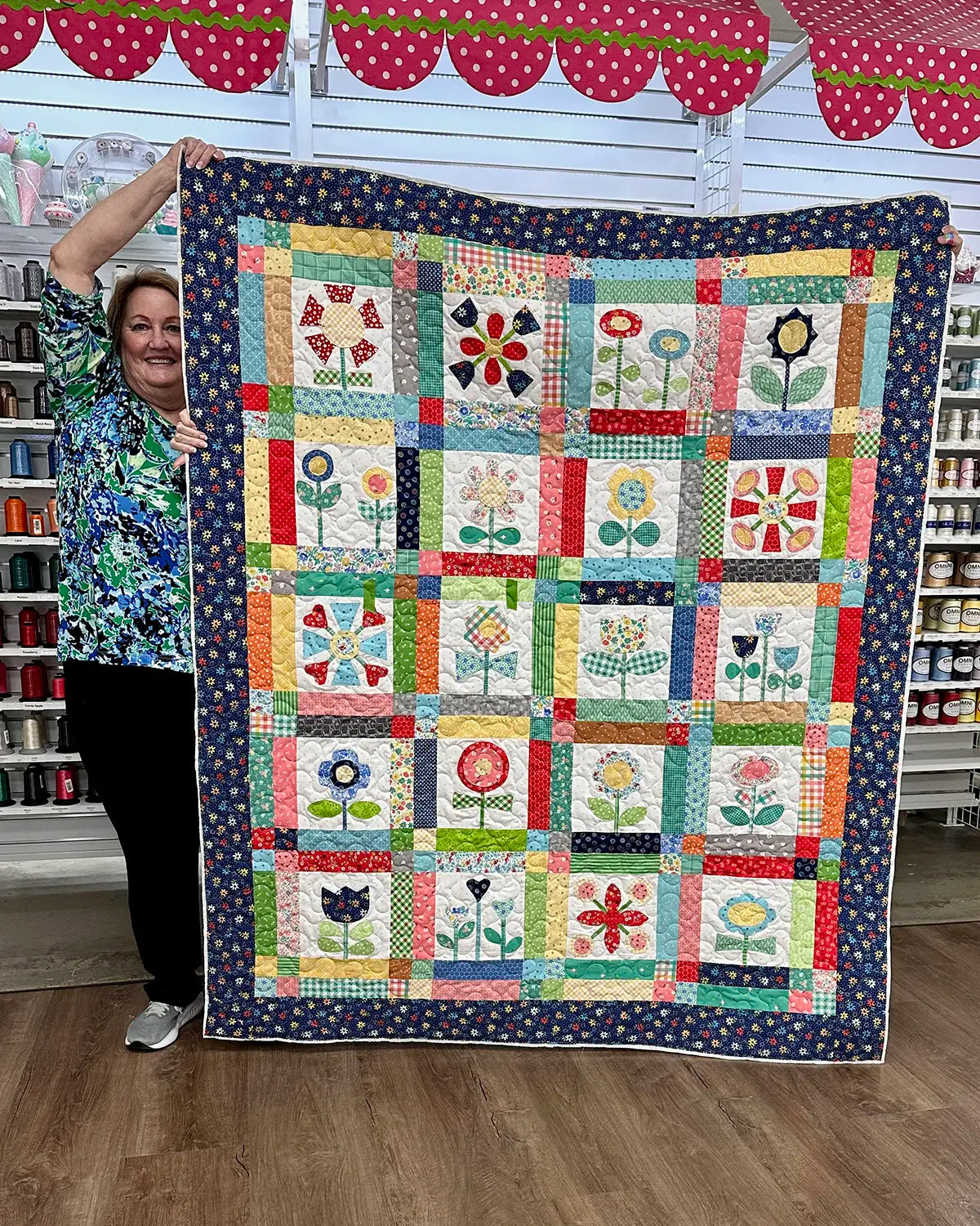 A smiling woman stands indoors holding up a colorful patchwork quilt decorated with various floral patterns. Shelves of thread and craft supplies are visible in the background.