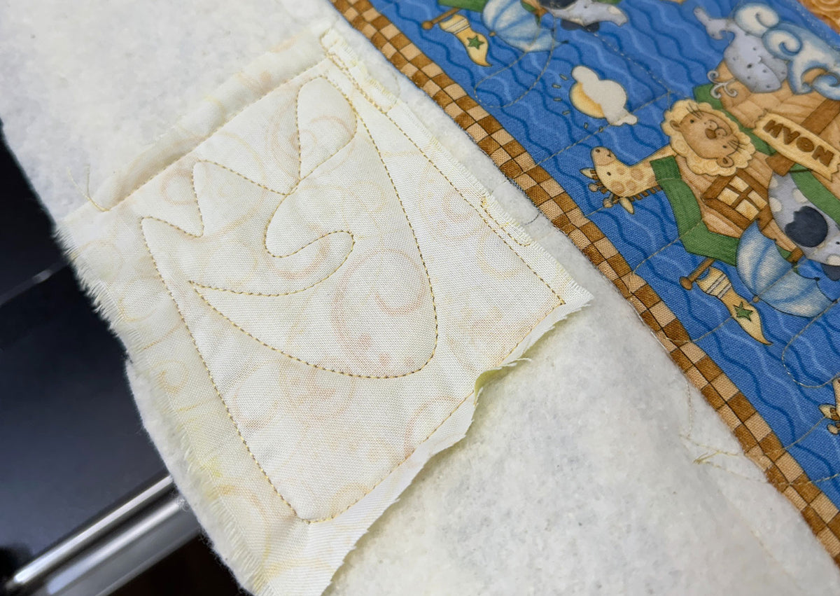 A close-up of a quilt in progress, showing a cream-colored fabric patch with a quilted chicken design next to a blue patch featuring cartoon animals and a brown checked border.
