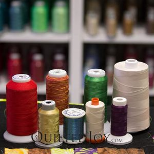 Several spools of thread in various colors and sizes are arranged on a surface, with blurred shelves of more thread spools in the background.